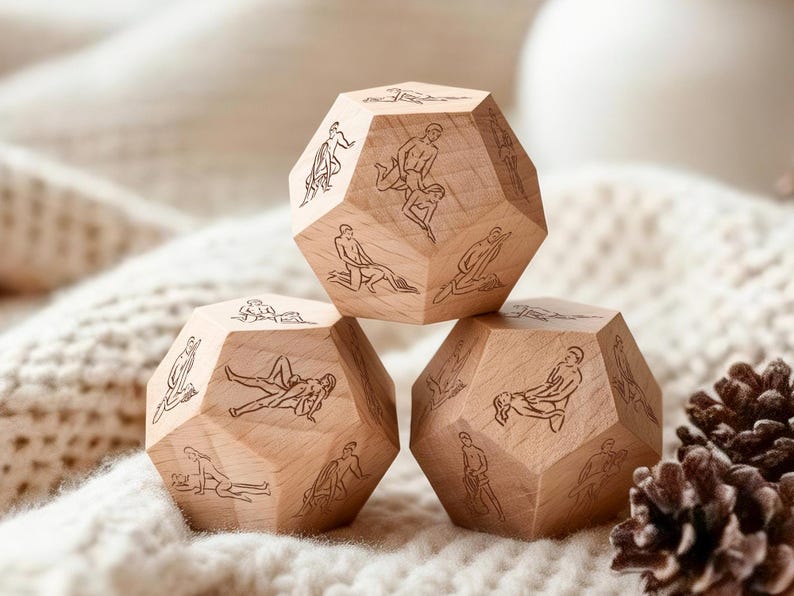 May include: Three wooden dodecahedron dice with etched illustrations of various sexual positions. The dice are made of light-colored wood and are arranged on a soft, white, textured surface. A pine cone is visible in the background.