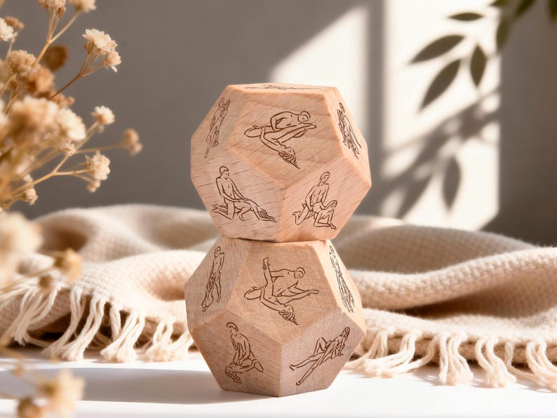 May include: Two wooden, multi-sided dice with etched illustrations of various sexual positions. The dice are stacked on top of each other, with a neutral-toned blanket in the background. Dried flowers are in the foreground.