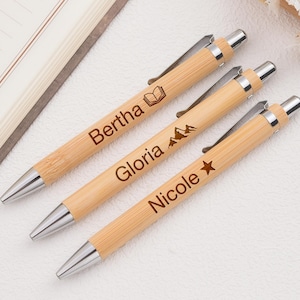 Puede incluir: Tres bolígrafos personalizados de bambú con detalles plateados. Cada bolígrafo está grabado con un nombre y un pequeño icono: un libro, montañas y una estrella. Los bolígrafos están sobre una superficie clara junto a un cuaderno.