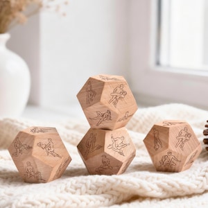 May include: Four wooden dice with engraved illustrations of various sexual positions. The dice are light brown and sit on a cream-colored knitted blanket. A white vase with dried flowers is in the background.