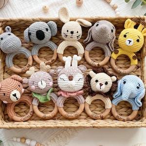 May include: A collection of handmade crochet baby rattles in a woven basket. The rattles feature various animal designs, including a koala, bunny, elephant, giraffe, bear, deer, and lion, with wooden ring handles. Each ring has a name engraved.
