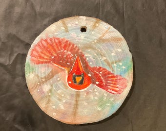 May include: A hand-painted wooden ornament featuring a red cardinal bird flying through a snowy winter scene. The background is painted in shades of blue, green, and white, with white dots representing snowflakes.