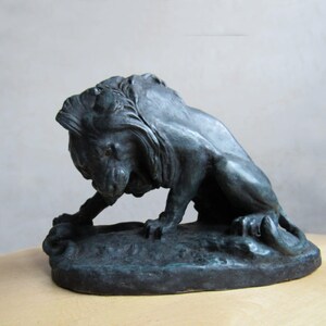 Könnte beinhalten: Eine dunkelgrüne Bronzeskulptur eines Löwen, der auf einem Sockel hockt. Der Löwe hat eine detaillierte Mähne und ist in einer realistischen Pose dargestellt.