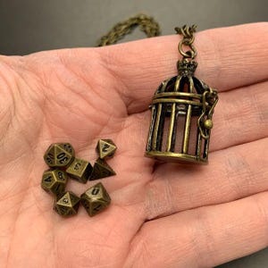 May include: A bronze-colored cage pendant necklace with a matching chain, next to a collection of bronze polyhedral dice. The cage is intricately detailed, and the dice have visible numbers. The items are displayed on a neutral background.
