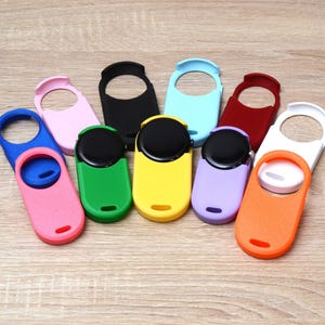 May include: A collection of eleven silicone protective covers for smartwatches, each featuring a circular cutout for the watch face and a small slot at the bottom. The covers are available in various solid colors including pink, blue, green, yellow, black, light blue, red, white, purple, and orange.