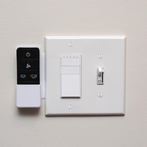 May include: A white wall plate with a light switch, a dimmer switch, and a remote control. The remote control is black with white buttons and icons. The light switch is in the on position.