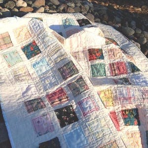 May include: A patchwork quilt featuring a grid of white fabric squares interspersed with various colorful patterned fabric squares in shades of blue, pink, red, and black. The quilt has visible stitching lines forming a square grid pattern throughout.