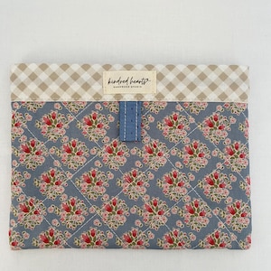 May include: A rectangular quilted pouch featuring a blue fabric with a repeating red and green floral pattern. The top edge has a beige and white gingham border with a central label reading Kindred Hearts Handmade Studio and a blue tab.