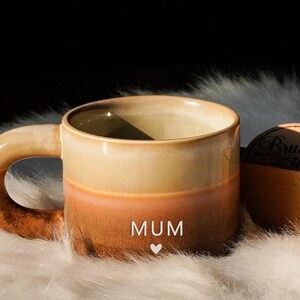 Puede incluir: Una taza de cerámica con un degradado de color, que va del beige al naranja y al marrón. La palabra "MUM" está impresa en la parte delantera con un símbolo de corazón. La taza tiene un asa grande y curvada y está sobre una superficie blanca y esponjosa.