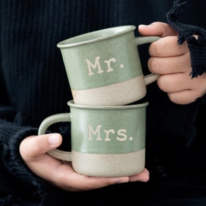 May include: Two stacked green ceramic mugs with a speckled, off-white base. The top mug reads "Mr." and the bottom mug reads "Mrs." in a simple, sans-serif font. The mugs are held in front of a dark background.
