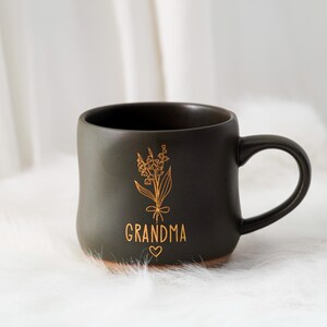以下が含まれることがあります： 金色の花の模様と「GRANDMA」の文字が金色で描かれた、濃い灰色のセラミックマグカップ。 マグカップは丸みを帯びた形状で、取っ手と茶色の底があります。 デザインにはハートのシンボルが含まれています。