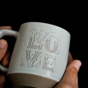 以下が含まれることがあります： 装飾的なフォントで「LOVE」の文字がエンボス加工された、水色のセラミックマグカップ。マグカップの底は濃い茶色です。黒い背景を背景に、人の手でマグカップが持たれています。