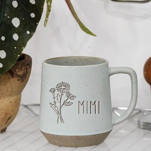 Puede incluir: Una taza de cerámica azul claro con base marrón, con un diseño floral y el nombre "MIMI". La taza está sobre una estantería de alambre, con una planta y un gancho de madera en el fondo.