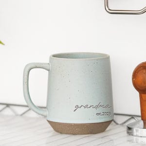 以下が含まれることがあります： 水色のセラミックマグカップ。まだら模様の仕上がりと茶色の底が特徴です。「grandma」の文字が筆記体で前面に書かれており、その下に「est. 2025」と書かれています。マグカップにはカーブしたハンドルが付いています。