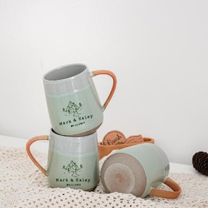 Puede incluir: Tres tazas de cerámica apiladas, cada una con un cuerpo verde claro, borde gris y asa naranja. Las tazas están personalizadas con un gráfico de montaña y los nombres "Mark & Haley" y la fecha "04.11.2025".