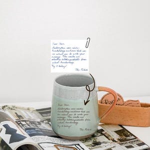 Puede incluir: Una taza de cerámica con un esmalte verde claro y blanco, con un asa color melocotón. La taza tiene texto escrito a mano y está colocada sobre una revista. También es visible un pequeño recipiente de madera con objetos redondos.