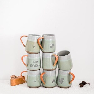Puede incluir: Una pila de tazas de cerámica hechas a mano en tonos verdes y grises, con asas naranjas. Algunas tazas presentan diseños de hojas o corazones, mientras que una muestra "CUSTOM TEXT/LOGO". También hay una caja de madera y piñas.