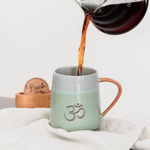 Puede incluir: Una taza de cerámica verde claro con el símbolo Om se está llenando con líquido oscuro de una jarra de vidrio. La taza tiene un borde gris y un asa de color cobre. Un posavasos de madera con la palabra "Brush" está en el fondo.