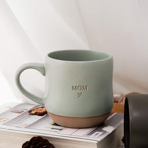 Peut inclure: Une tasse en céramique vert clair avec le mot "MOM" et un symbole de cœur. La tasse a une forme arrondie, une base marron et une anse incurvée. Elle est posée sur une pile de livres.