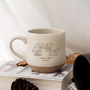 Könnte beinhalten: Becher aus gesprenkelter Keramik mit braunem Boden und Henkel. Der Becher zeigt eine Strichzeichnung von zwei Elefanten, die sich mit den Rüsseln berühren, mit dem Text "Lucy & Bob 06.06.2012" darunter. Der Becher steht auf einem Buch.