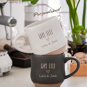 Puede incluir: Dos tazas de cerámica personalizadas apiladas. Una es blanca, la otra gris oscuro. Ambas tazas tienen una base marrón, un asa y el texto "Lucy & Jack" con un símbolo de corazón. Las tazas también presentan un diseño de marcas de conteo.