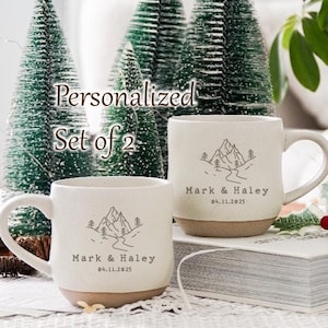 May include: Set of two personalised white ceramic mugs with a mountain and river design. The mugs have the text "Mark & Haley 04.11.2025" on them.