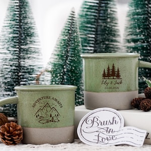 Puede incluir: Dos tazas de cerámica con un esmalte verde claro moteado en la mitad superior y una base beige mate. Una taza presenta una montaña, una tienda de campaña y el texto "ADVENTURE AWAITS". La otra muestra un gráfico de bosque con "Lily & Jack 02.06.2026".