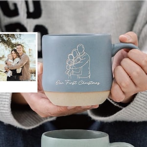 Puede incluir: Una taza de cerámica azul con base beige, con una ilustración familiar y el texto "Our First Christmas". La taza está sostenida en las manos de alguien, con una foto enmarcada de una familia en el fondo. La taza tiene un asa curva.