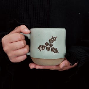 May include: A light blue ceramic mug with a brown base and a handle. The mug features a brown holly design. The mug is held in a person's hands against a dark background. The mug is a standard size.