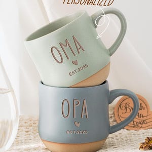 May include: Two ceramic mugs with personalized text. The top mug is light green with the word "OMA" and "EST. 2025". The bottom mug is gray with the word "OPA" and "EST. 2025".