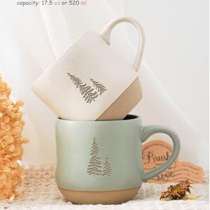 May include: Two ceramic mugs with a tree design. The top mug is white with a brown rim and the bottom mug is green with a brown rim. Both mugs have a capacity of 17.5 oz or 520 ml.