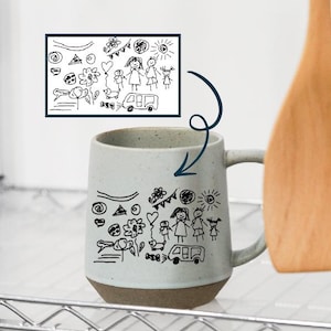 Pode incluir: Uma caneca de cerâmica salpicada com base castanha e alça branca. A caneca é decorada com desenhos a traço preto de uma família, flores e um veículo. O desenho é repetido numa imagem emoldurada.