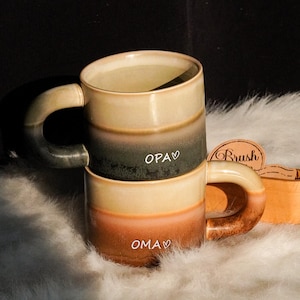 Puede incluir: Dos tazas de cerámica apiladas con una combinación de colores degradados de beige, marrón y verde. Las tazas tienen asas grandes y curvas. La palabra "OMA" está escrita en la taza inferior y "OPA" en la superior.