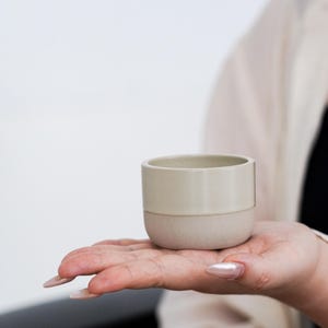 Può includere: Una piccola tazza in ceramica bicolore. La metà superiore è beige chiaro e la metà inferiore è beige leggermente più scuro e strutturato. La tazza è tenuta nella mano di una persona, con uno sfondo bianco.