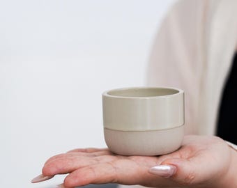Peut inclure: Une petite tasse en céramique bicolore. La moitié supérieure est beige clair et la moitié inférieure est beige légèrement plus foncé et texturé. La tasse est tenue dans la main d'une personne, sur fond blanc uni.