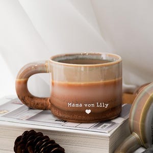 以下が含まれることがあります： ベージュから茶色へのグラデーションカラーのセラミックマグ。 大きなハンドルがあり、「Mama von Lily」の文字とハートのシンボルが付いています。 マグカップは、松ぼっくりの隣の本の上に置かれています。