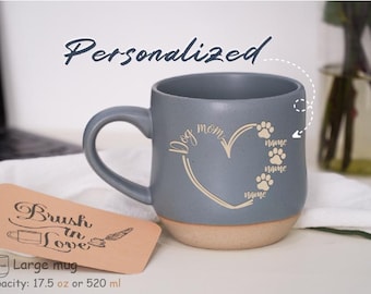 Puede incluir: Una taza de cerámica gris con un diseño de corazón y huellas de patas. La taza dice "Dog Mom" y tiene espacio para tres nombres de perros. La taza tiene una capacidad de 520 ml.
