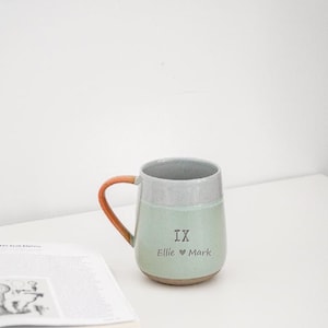 Puede incluir: Una taza de cerámica con cuerpo verde claro, borde gris y asa marrón y naranja. La taza tiene el texto "IX Ellie ♥ Mark" impreso en la parte delantera. La taza está sobre una superficie blanca.