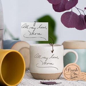 Könnte beinhalten: Eine weiße Keramiktasse mit braunem Boden, auf der handschriftlich "All my love, Mom" steht. Die Tasse wird von anderen Keramiktassen in Gelb und Blau umgeben, mit lila Blättern im Hintergrund.