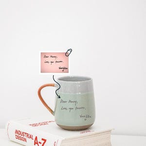 Puede incluir: Una taza de cerámica de dos tonos con base verde claro y parte superior gris, con asa marrón. La taza tiene texto escrito a mano que dice "Dear Henry: Love you forever. Yours, Elise." Una nota con el mismo texto está sujeta con un clip.