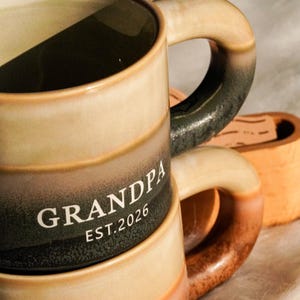 May include: A ceramic mug with a gradient design in shades of beige, brown, and black. The word "GRANDPA" and the year "EST.2026" are printed in white. The mug has a curved handle and is part of a set.