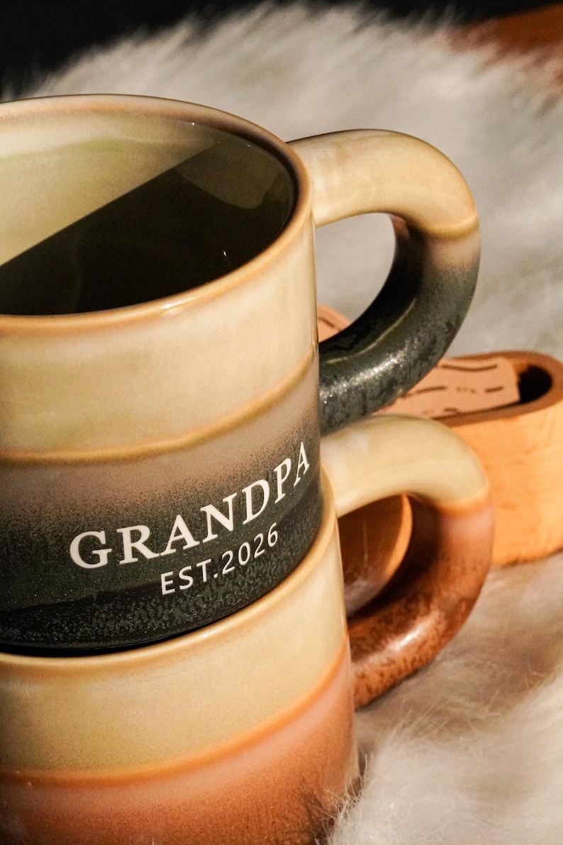 May include: A ceramic mug with a gradient design in shades of beige, brown, and black. The word "GRANDPA" and the year "EST.2026" are printed in white. The mug has a curved handle and is part of a set.