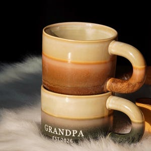 May include: Two stacked ceramic mugs with a gradient color scheme. The mugs transition from a light beige at the top and bottom to a brown and orange middle section. The word "GRANDPA" and "EST 2026" are printed on the bottom mug.