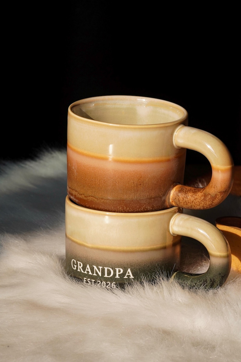 May include: Two stacked ceramic mugs with a gradient color scheme. The mugs transition from a light beige at the top and bottom to a brown and orange middle section. The word "GRANDPA" and "EST 2026" are printed on the bottom mug.