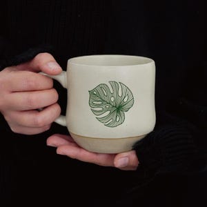 Op de afbeelding: Crèmekleurige keramische mok met een groen monstera blad ontwerp. De mok heeft een afgeronde vorm, een lichtbruine basis en een handvat. Het bladontwerp is gecentreerd op de voorkant van de mok.