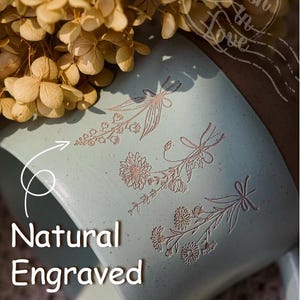May include: A pale blue ceramic mug featuring engraved floral patterns and the words "Natural Engraved". The mug has a brown rim and a handle. Dried flowers are in the background.