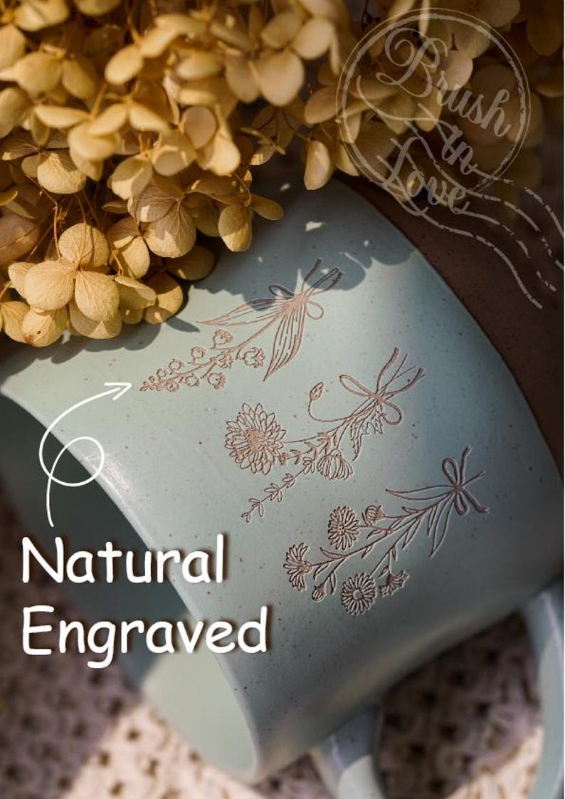 May include: A pale blue ceramic mug featuring engraved floral patterns and the words "Natural Engraved". The mug has a brown rim and a handle. Dried flowers are in the background.