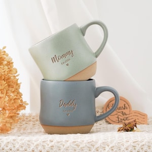 Può includere: Due tazze in ceramica impilate. La tazza superiore è verde chiaro e dice "Mommy" con "Est 2026" e un cuore. La tazza inferiore è grigio-blu e dice "Daddy" con "Est 2026" e un cuore. Entrambe le tazze hanno una base marrone.