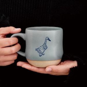 May include: A gray ceramic mug with a duck illustration. The mug has a light gray upper half and a tan lower half. The duck is navy blue. The mug is held in a person's hands against a black background.