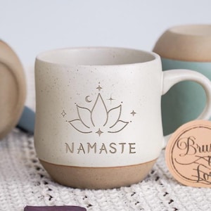Op de afbeelding: Witte keramische mok met een gespikkelde afwerking. De mok heeft een bruine basis en een handvat. De mok heeft een ontwerp met een lotusbloem met sterren en een halve maan. De tekst "NAMASTE" is onder het ontwerp gedrukt. De mok staat op een wit gehaakt oppervlak.